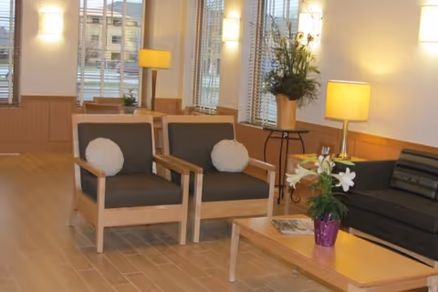 A well-lit living room area with two wooden armchairs featuring dark cushions and round white pillows, a wooden coffee table with a purple vase holding white flowers, a dark sofa, two table lamps with yellow shades, and large windows with blinds letting in natural light.