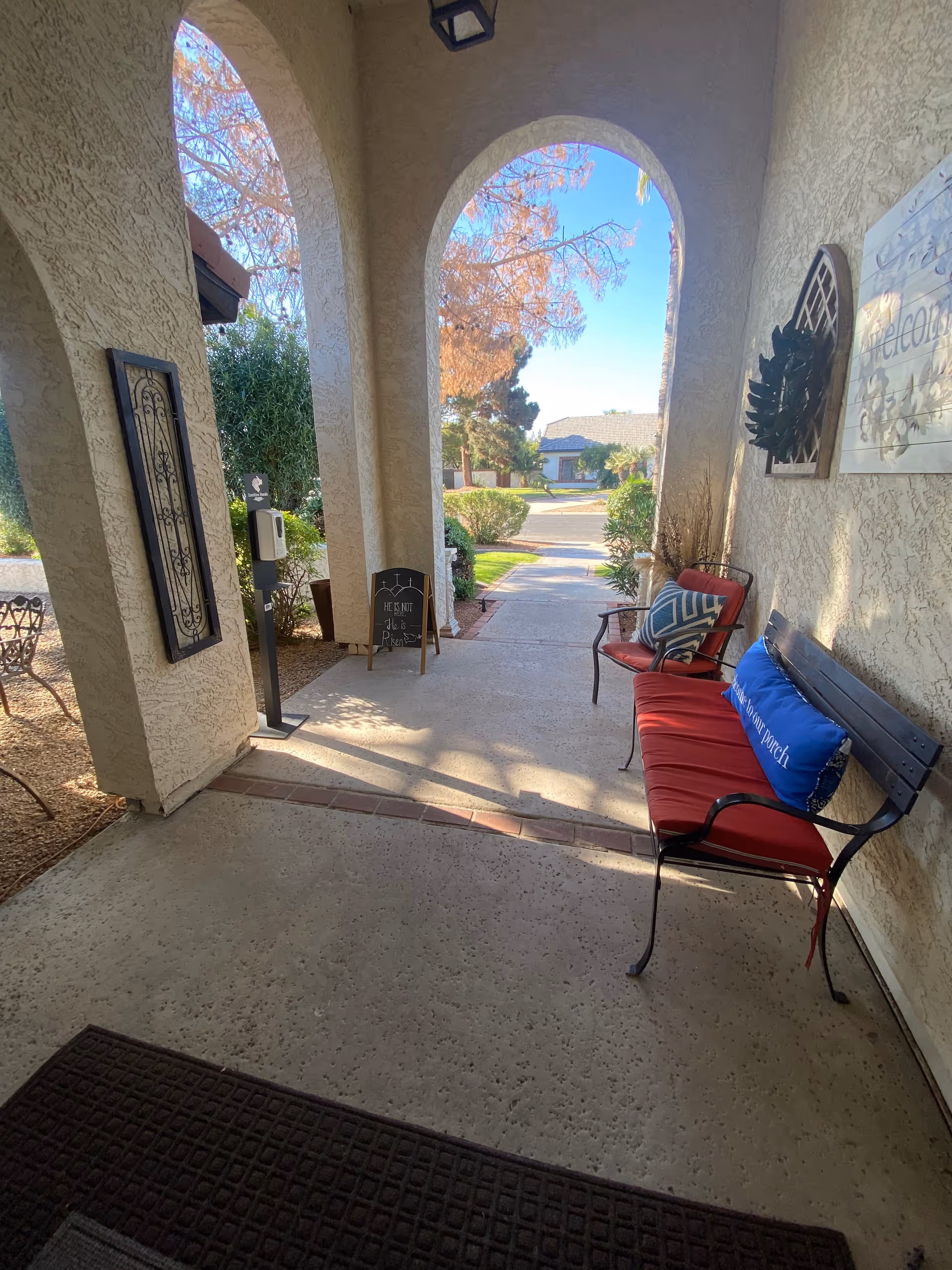 Covered outdoor porch area with arched openings, a bench with red cushions and pillows, a chair with a cushion, decorative wall hangings, a small chalkboard sign, and a view of a sidewalk leading to a residential street with trees and houses.