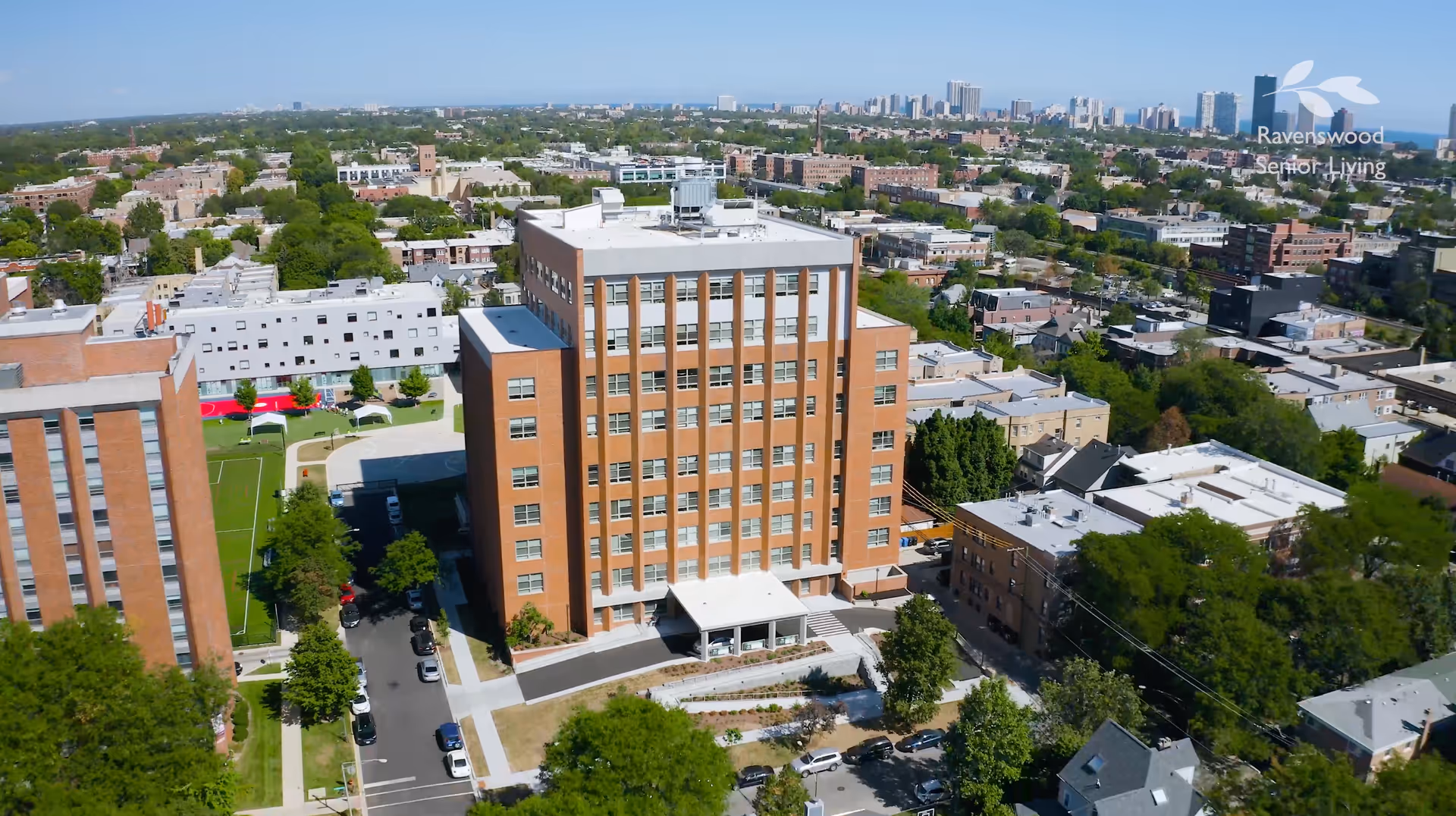 Aerial view of a multi-story brick building surrounded by trees and other buildings in an urban area under a clear blue sky. The building has a white entrance canopy and is part of a larger residential or supportive living complex. The city skyline is visible in the distance.