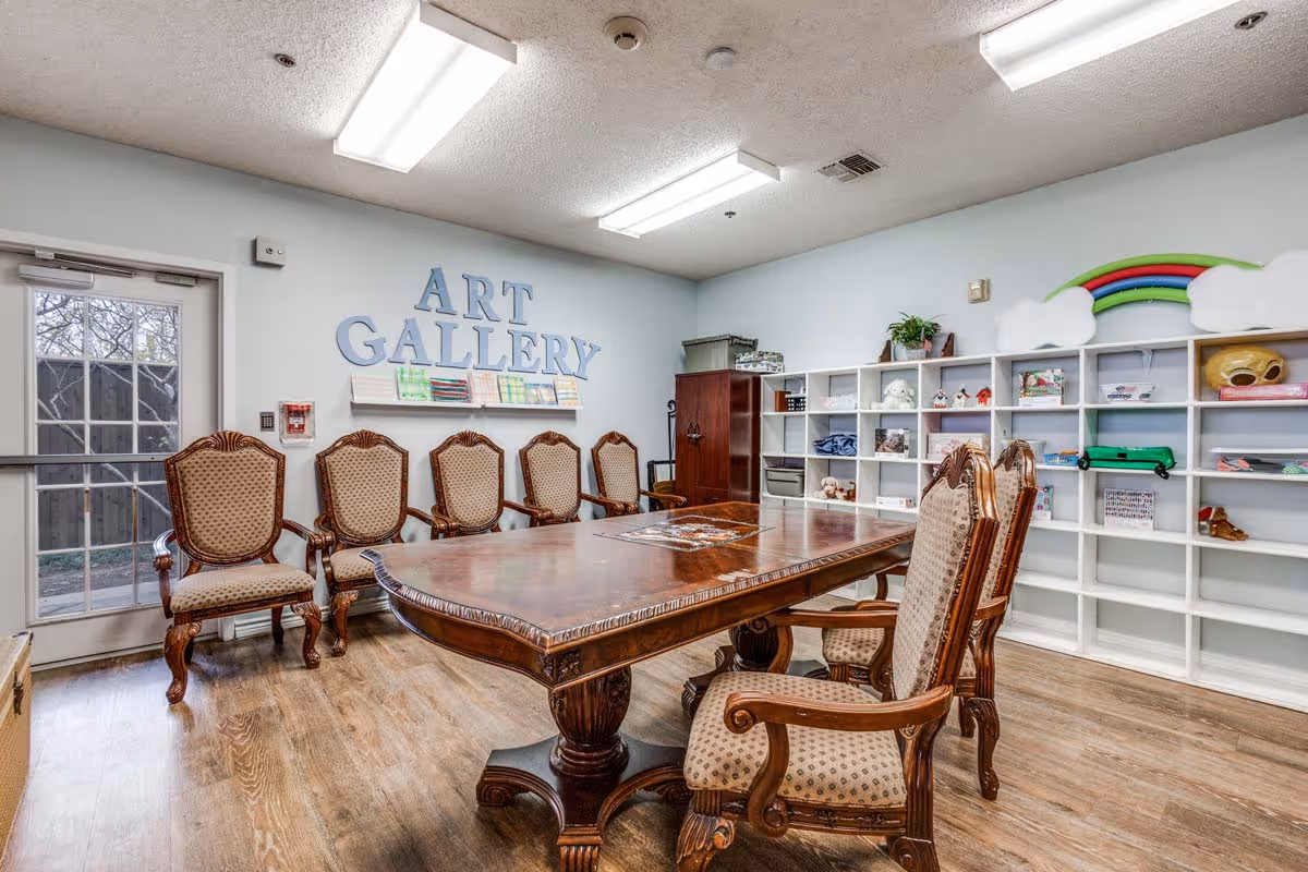 A bright art activity room with a large wooden table surrounded by upholstered chairs and wall shelving under an 'ART GALLERY' sign.
