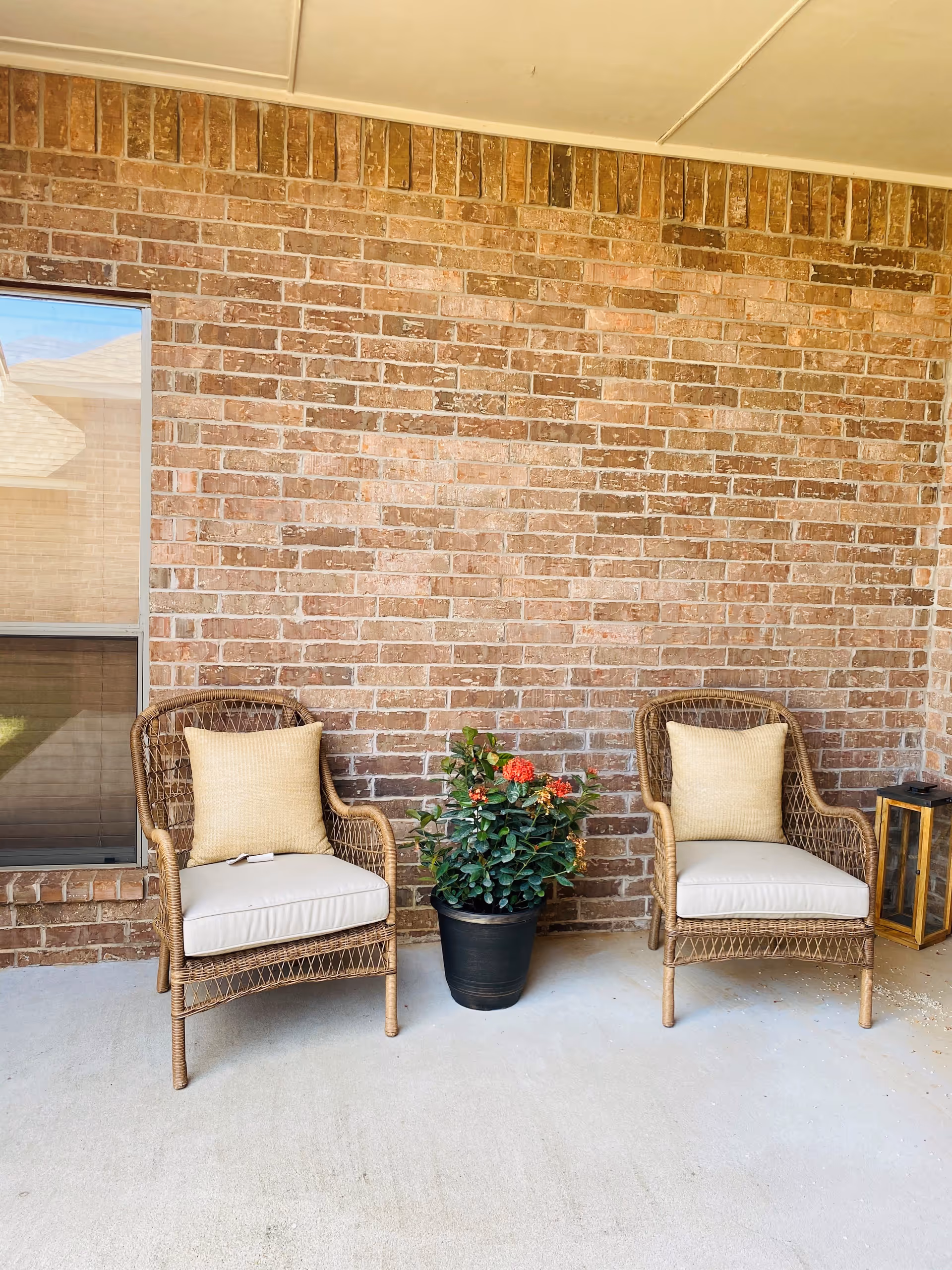 Two wicker chairs with beige cushions and pillows are placed on a covered patio against a brick wall. Between the chairs is a black pot with a green plant that has red flowers. A window is visible on the left side, and a lantern-style decorative item is on the right side near one of the chairs.