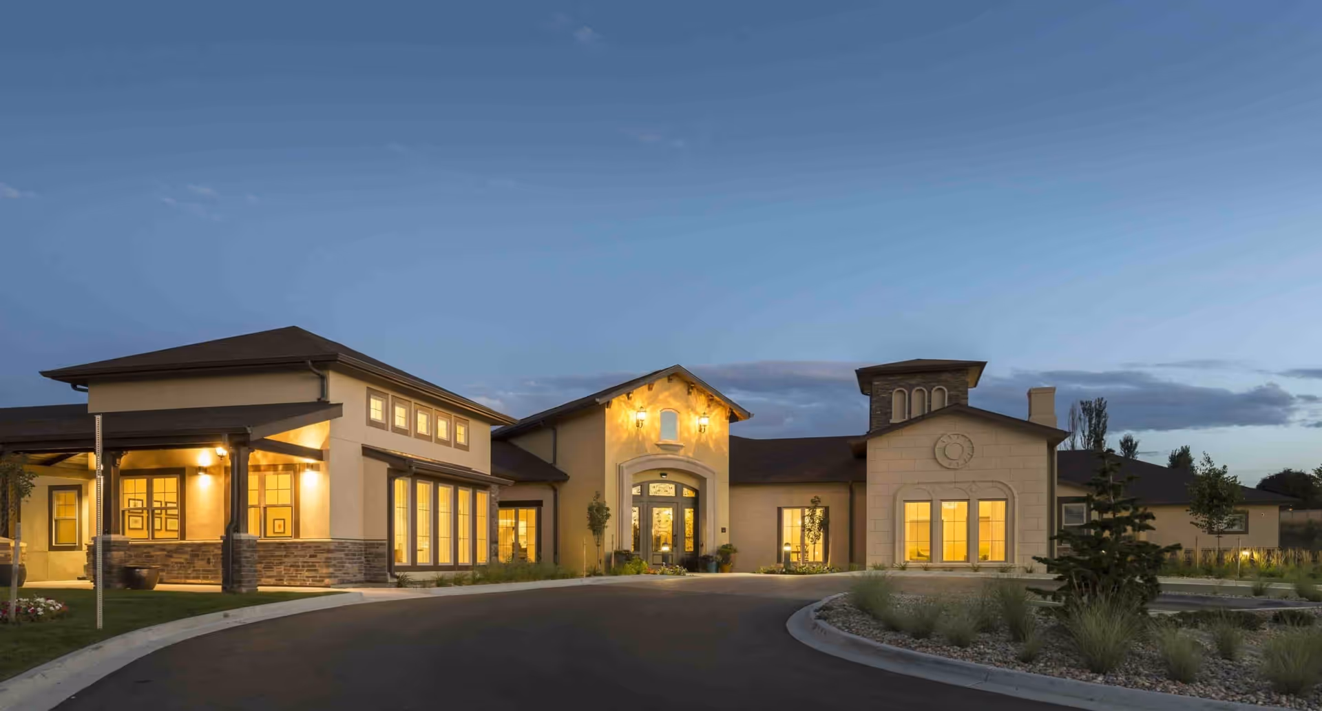 Exterior view of Villagio at Broomfield senior living facility at dusk, showing a well-lit building with multiple windows, a central entrance with decorative lighting, and landscaped surroundings including a curved driveway and greenery.