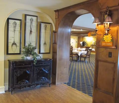 Interior view of a senior living facility showing a wooden cabinet with decorative framed artwork of utensils above it, a plant on top of the cabinet, and an arched wooden doorway leading into a dining area with tables and chairs. A wall sconce with two lights and a small decorative sign that says 'GIVE THANKS' is mounted on the wooden arch.