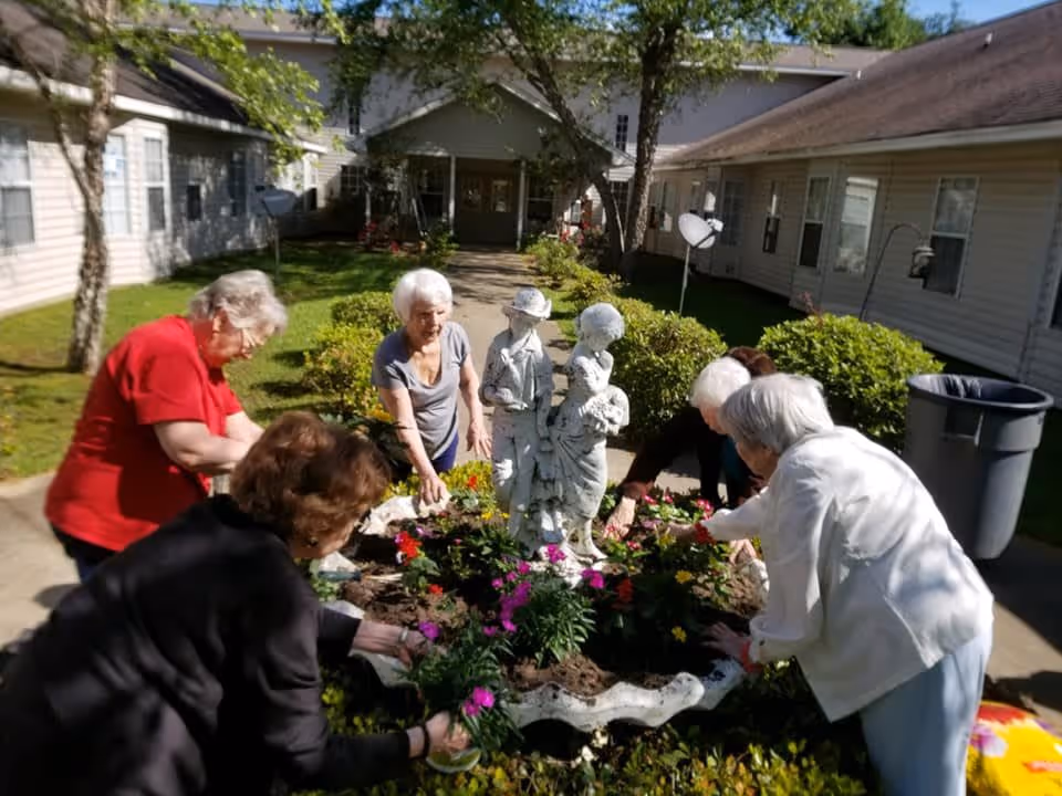 A group of elderly women gardening together around a flower bed with a decorative statue of children in the center, outside a residential facility with beige siding and green bushes.
