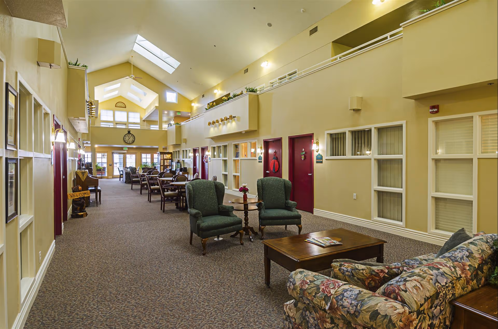 Spacious interior common area of Cambridge Court Apartments featuring high vaulted ceilings with skylights, multiple seating arrangements including floral-patterned sofa, green armchairs, wooden tables, and dining chairs. The walls are painted light yellow with red doors and windows, and there is a large clock on the far wall above the exit doors.