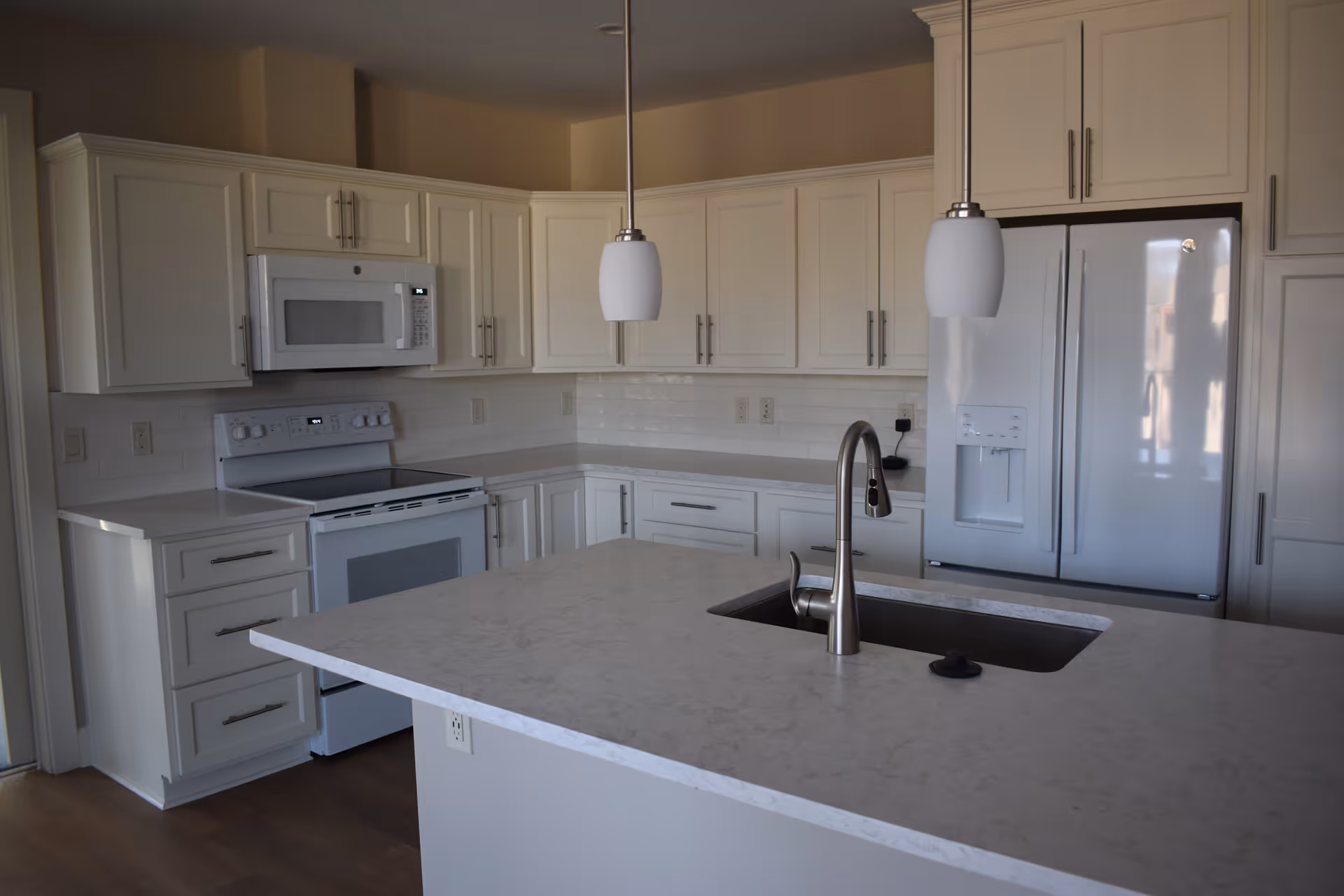 Modern kitchen with white cabinets, a white stove with an overhead microwave, a large white refrigerator, and a central island with a sink and a silver faucet. Two pendant lights hang above the island.
