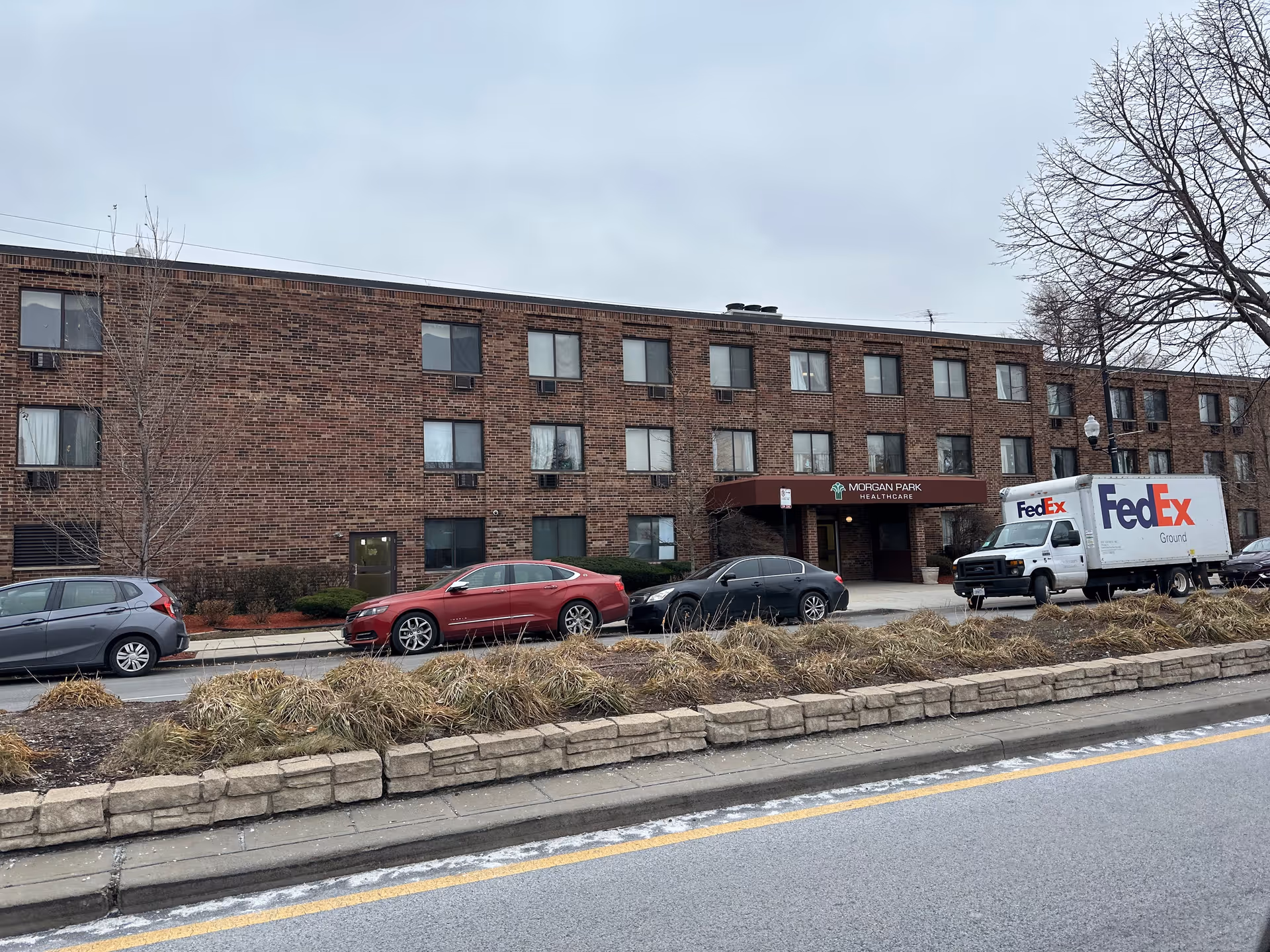 Exterior view of a three-story brick building with multiple windows, a maroon awning labeled 'Morgan Park Healthcare' above the entrance, several parked cars including a FedEx truck, and leafless trees on a cloudy day.