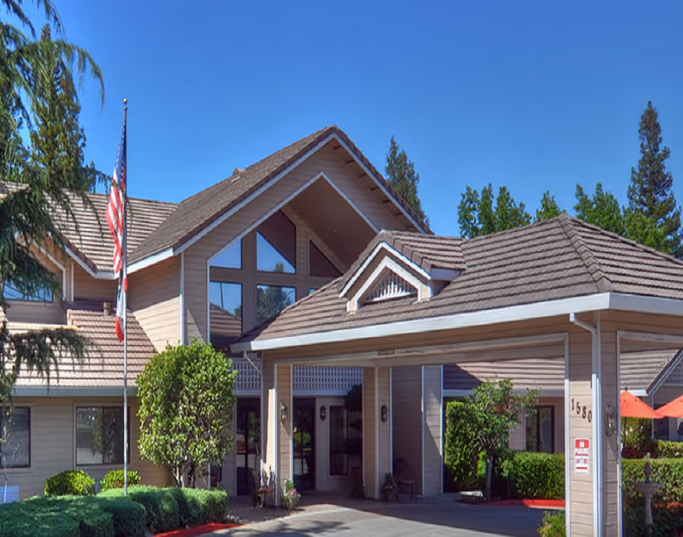 Exterior view of The Kensington at Walnut Creek senior living facility showing the entrance with a covered driveway, beige siding, large windows, and a flagpole with the American flag. The building is surrounded by green bushes and trees under a clear blue sky.