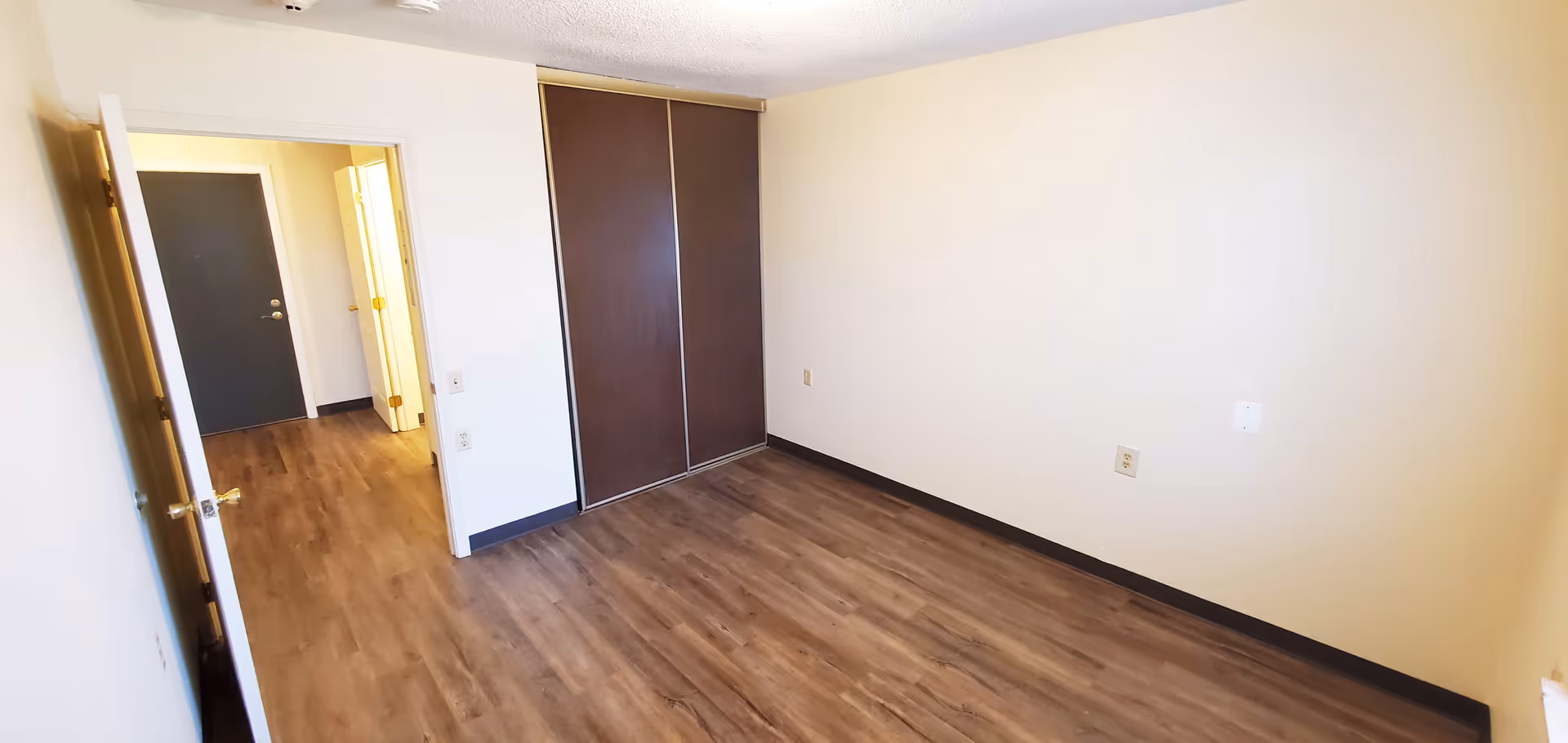 Empty room with wooden flooring, beige walls, a closed closet with dark sliding doors, and an open door leading to a hallway with a dark entrance door and another door visible inside.