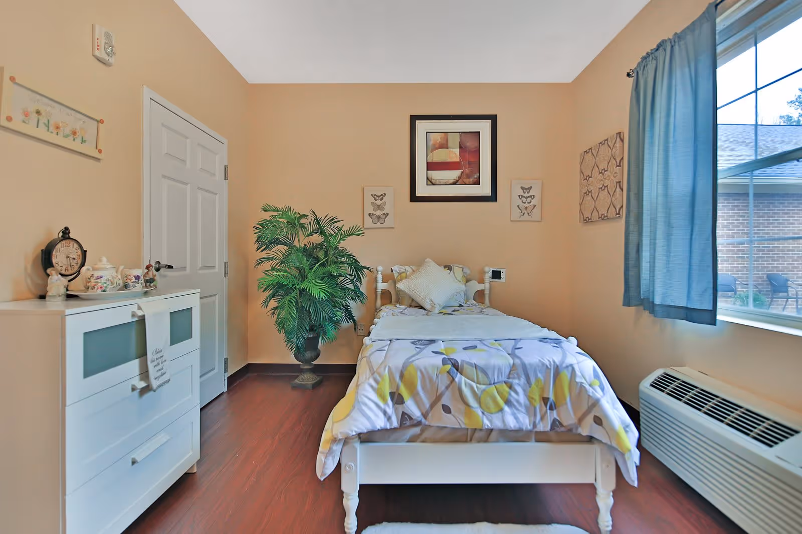 A tidy single bedroom with a twin bed, patterned bedding, white dresser, potted plant and a window with blue curtains.