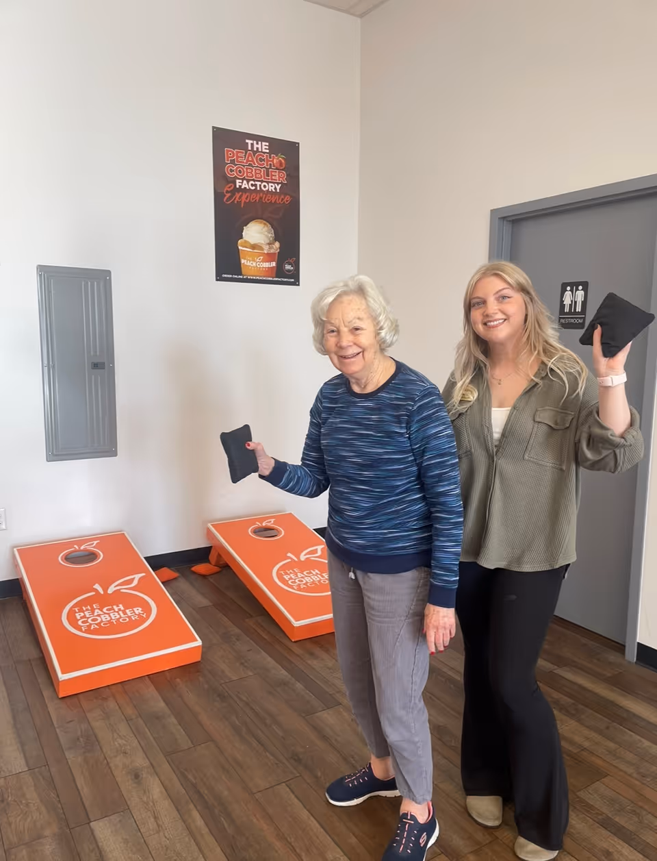 An elderly woman and a younger woman smiling and holding bean bags, standing in front of two orange cornhole boards with 'The Peach Cobbler Factory' logo in a room with wooden flooring and a restroom door in the background.