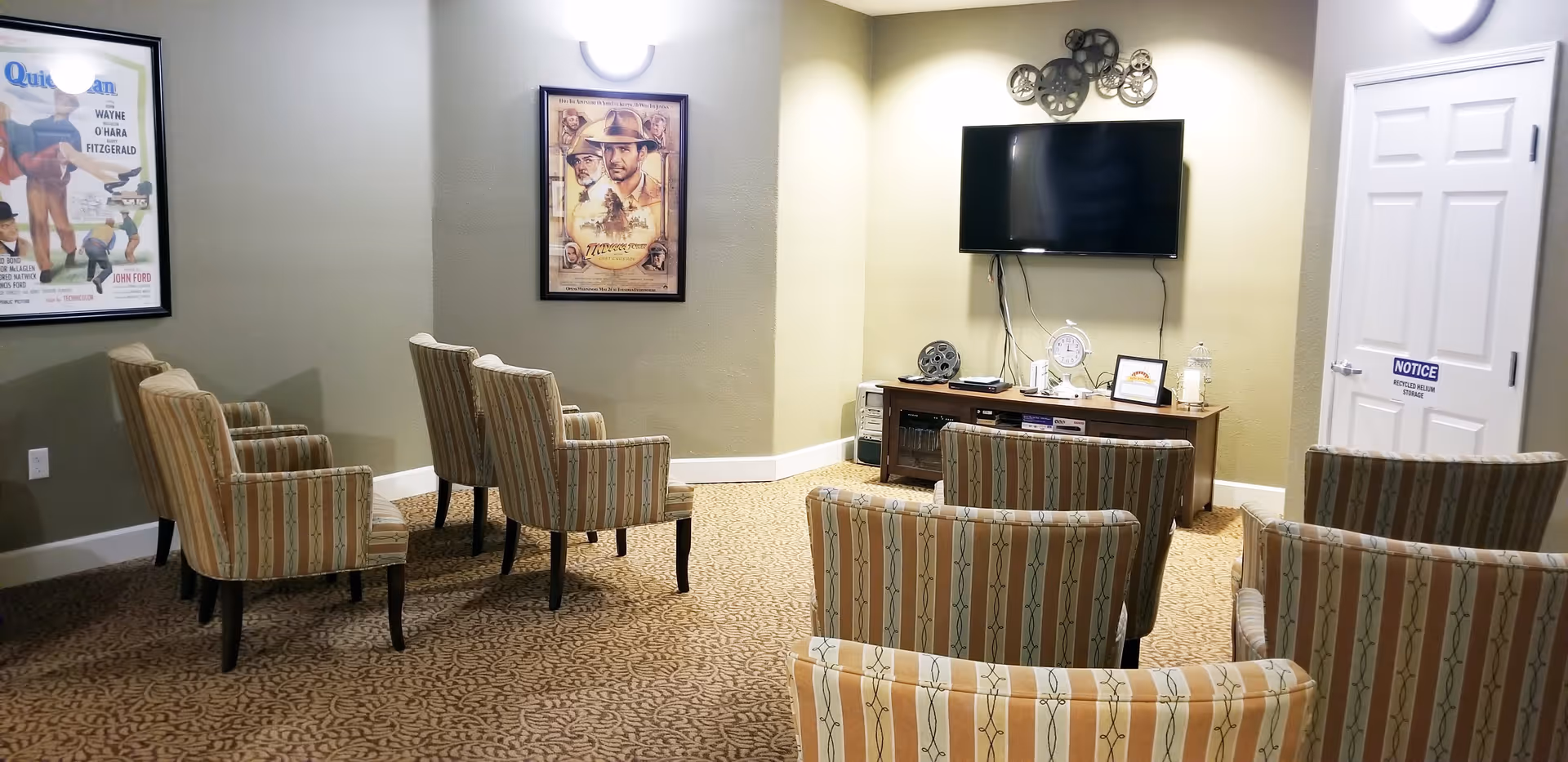 A small media room with eight striped upholstered chairs arranged in rows facing a wall-mounted flat screen TV. Below the TV is a wooden console table with decorative items including a clock and film reels. The walls are painted light green and beige, and there are two framed vintage movie posters on the walls. A white door with a notice sign is visible on the right side.