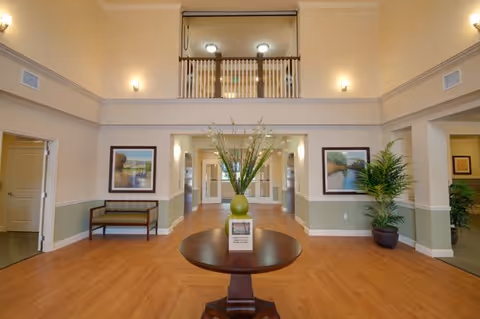 A spacious and well-lit interior common area with high ceilings and a wooden floor. In the center, there is a round wooden table with a green vase holding tall decorative plants. On the walls, there are framed landscape paintings and wall-mounted lights. There is a bench on the left side and potted plants on the right side. The area has doorways leading to other rooms and a balcony railing visible on the upper level.