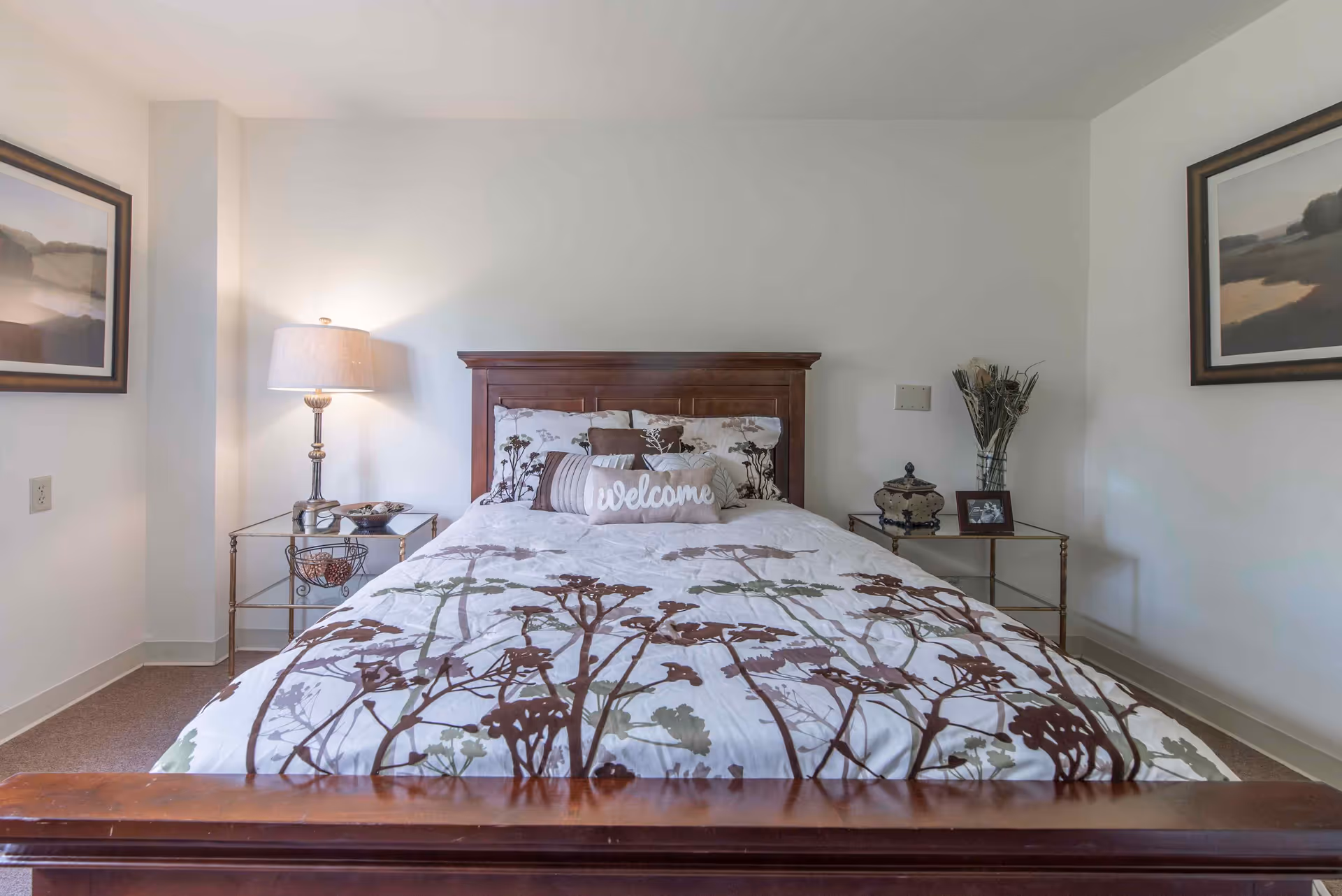 A neatly made bed with a wooden headboard and floral patterned bedding in a simple bedroom. There is a 'Welcome' pillow on the bed. On each side of the bed are glass-top nightstands; the left one has a lamp and decorative items, and the right one has a vase with dried flowers, a decorative container, and a framed photo. Two framed landscape paintings hang on the white walls.