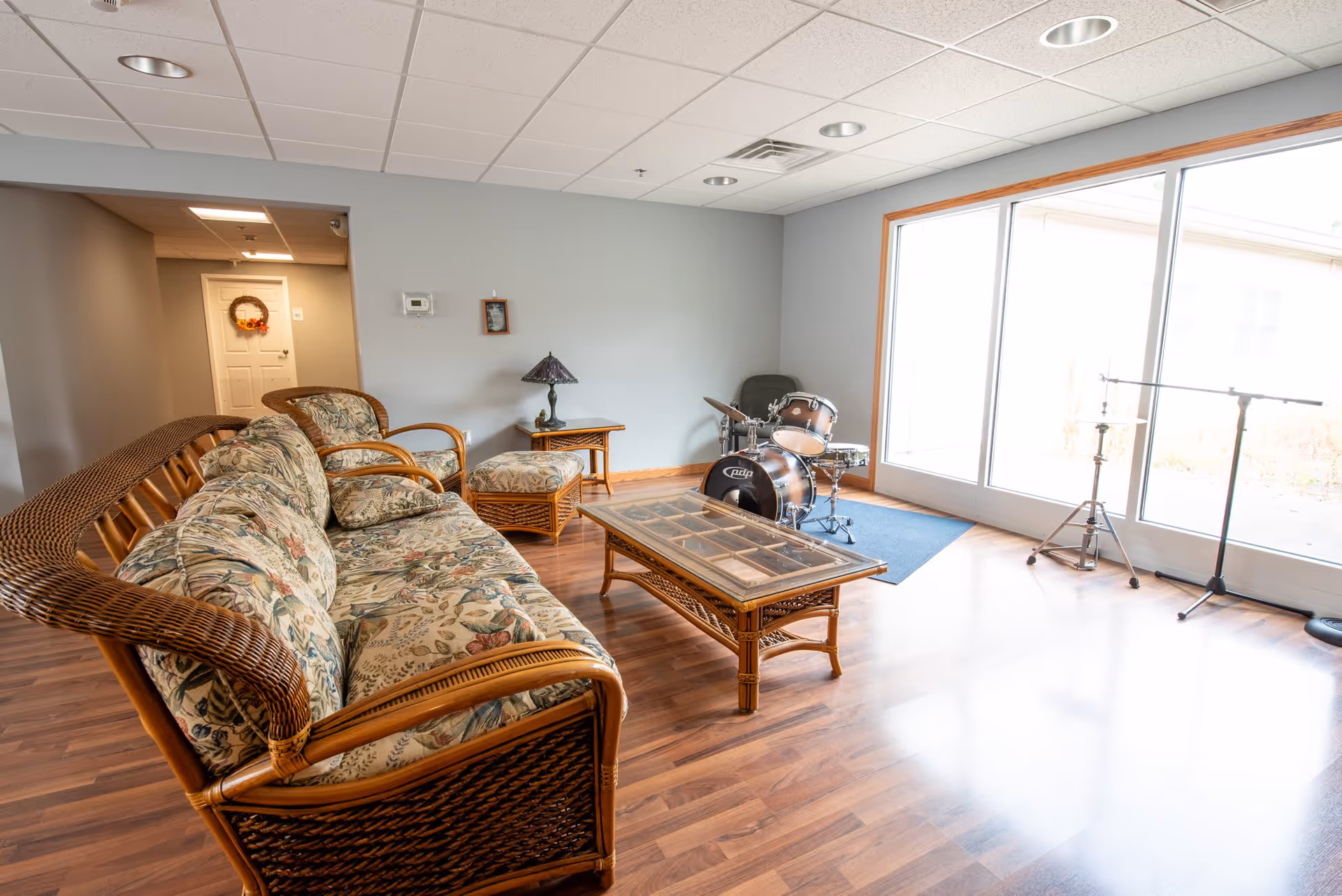 A cozy living room area with wicker furniture including a floral-patterned sofa, armchair, ottoman, and a glass-top coffee table. There is a small side table with a decorative lamp, a drum set on a blue rug near large sliding glass doors letting in natural light, and a door decorated with a wreath in the background.