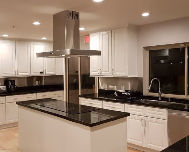 A modern communal kitchen with a central island, stainless steel range hood, black countertops, and white cabinets.