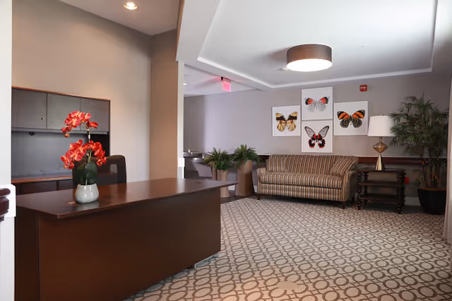 A reception area with a dark wooden desk adorned with a vase of red flowers. Behind the desk is a gray wall with cabinets. To the right, there is a seating area with a striped couch, a side table with a lamp, and several framed butterfly artworks on the wall. The floor is carpeted with a geometric pattern, and there are potted plants adding greenery to the space.