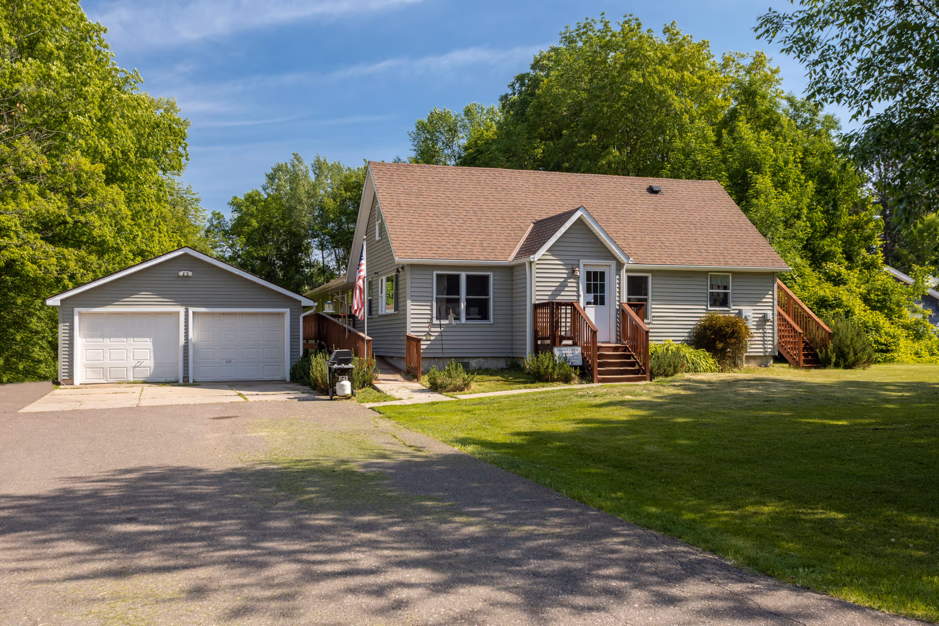 A single-story house with beige siding and a brown shingled roof, featuring a small front porch with wooden steps and railings. There is a detached two-car garage to the left of the house. The house is surrounded by green grass and trees under a clear blue sky.