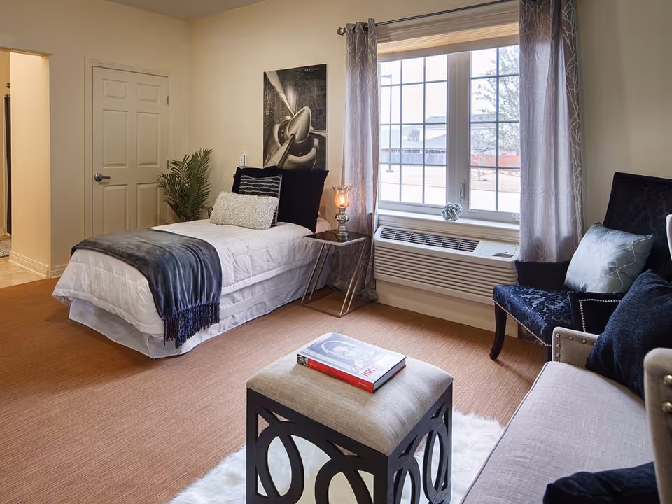 A cozy bedroom in a senior living facility featuring a single bed with white bedding and a dark throw blanket, a small side table with a lamp, a large window with curtains, a cushioned ottoman with a book on top, and comfortable seating including a chair and a sofa with pillows.