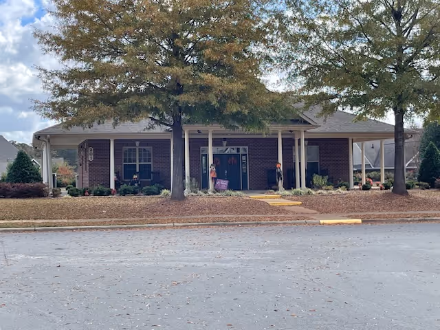 Brick single-story senior living facility entrance with a covered porch, two large trees in front, and a paved driveway.