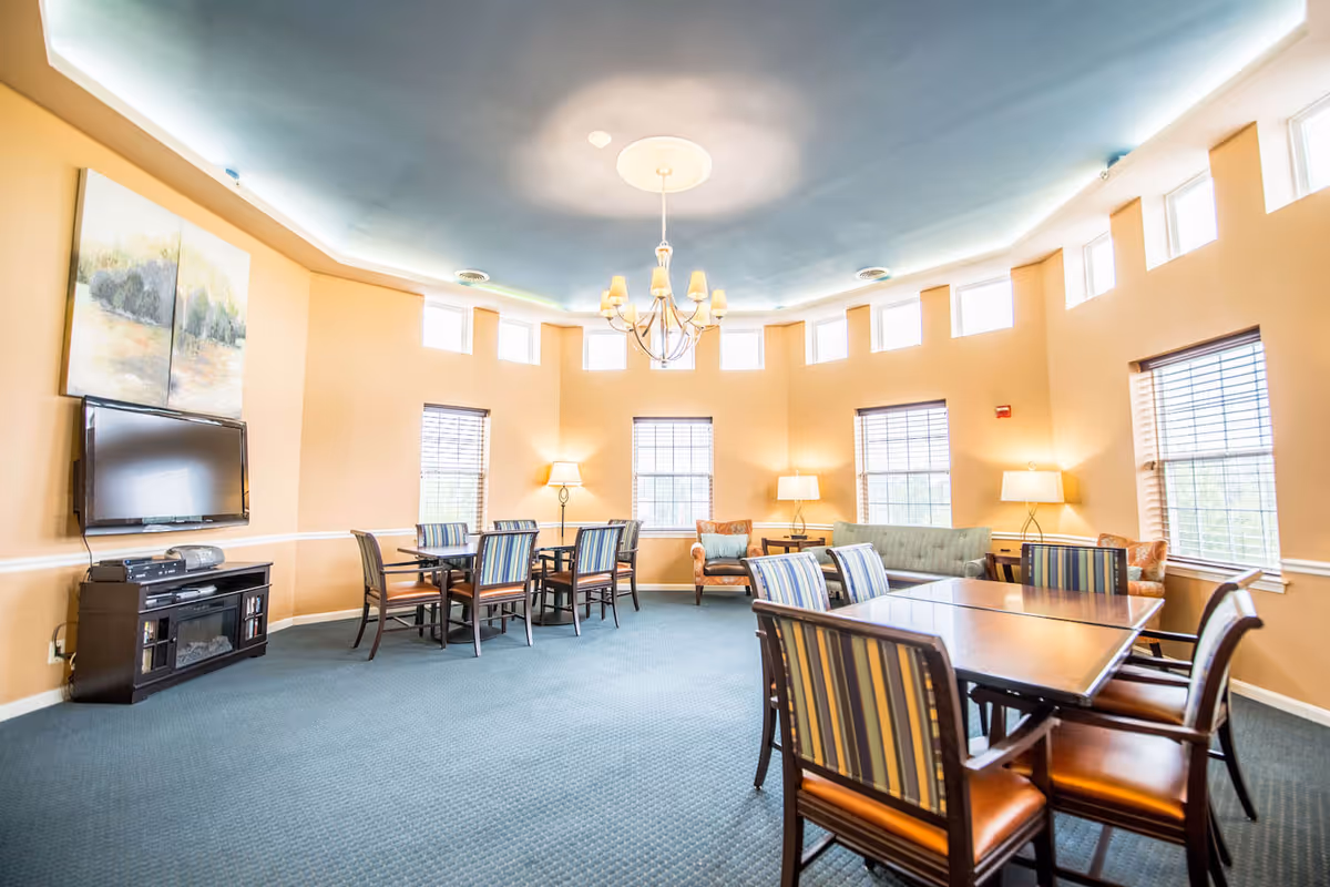 Well-lit communal room with dining tables and striped chairs, a TV, sofas and lamps beneath a decorative ceiling.