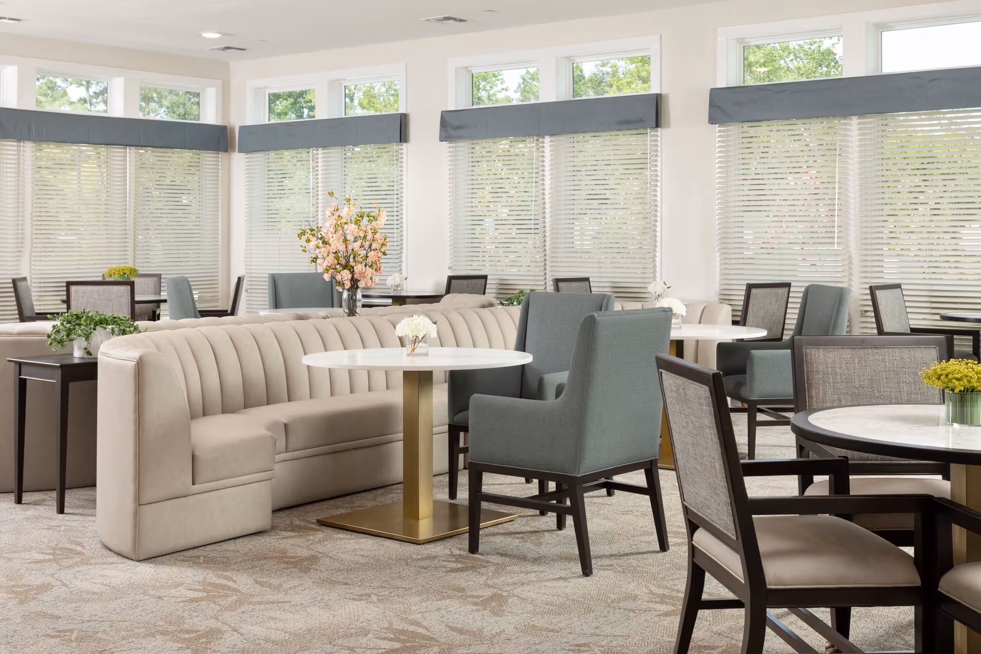 Bright dining/lounge area with upholstered banquette seating, round tables, assorted chairs, and large windows with blinds.
