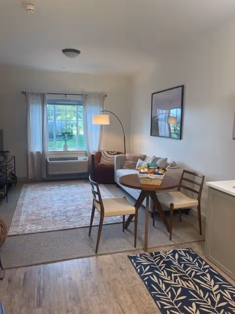 A cozy living room area with a round wooden table and two chairs in the foreground. Behind the table is a small sofa with cushions and a floor lamp with an arched arm. A window with sheer white curtains lets in natural light, and a patterned rug covers part of the floor. The walls are light-colored, and a framed picture hangs above the sofa.