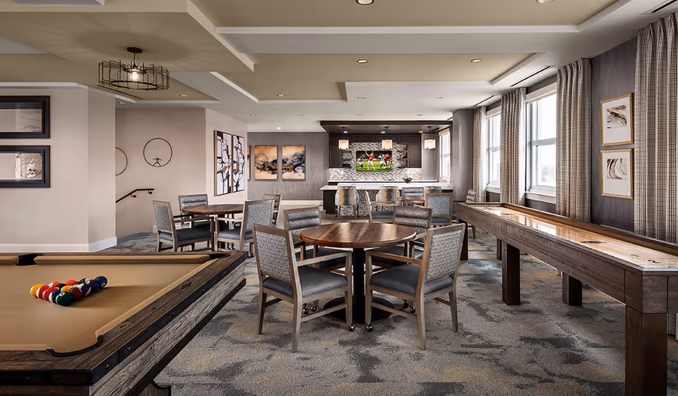 A spacious common area with a pool table on the left, round wooden tables with chairs in the center, and a shuffleboard table on the right. The back wall features a kitchen area with a TV mounted above a stone backsplash, pendant lights, and bar stools. Large windows with curtains allow natural light to fill the room. The walls are decorated with framed artwork.