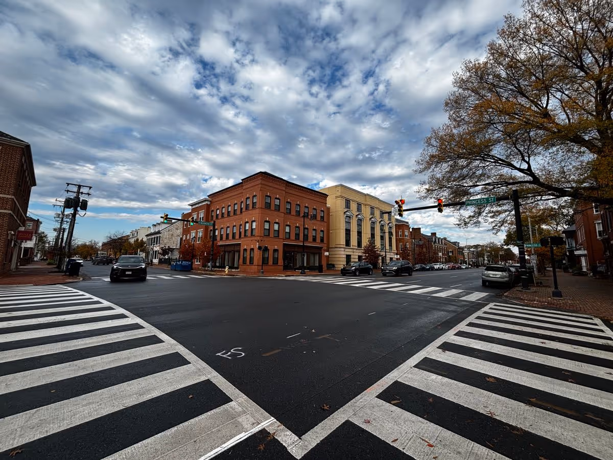 Street-level view of a crosswalk and intersection flanked by brick commercial buildings, cars, trees, and a cloudy sky.