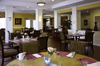 A spacious dining room with multiple tables set for meals, featuring chairs with patterned upholstery, place settings with napkins, cups, and small flower arrangements. The room has soft lighting, white columns, and walls painted in light yellow and white tones.