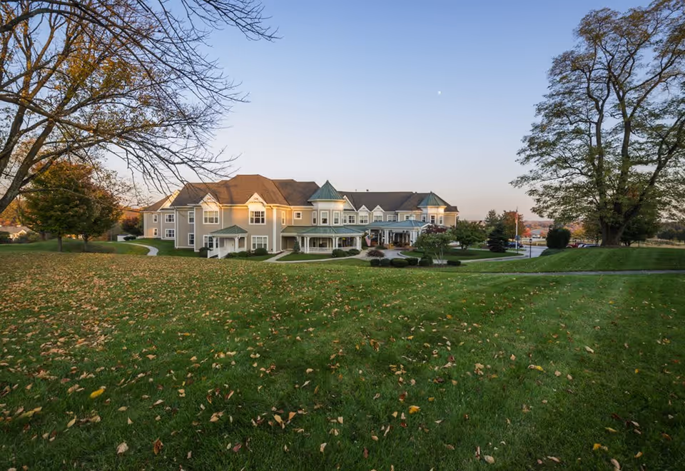 Exterior view of a large senior living facility building named Sunrise of Westtown, surrounded by green lawns with scattered autumn leaves and trees under a clear sky at dusk.