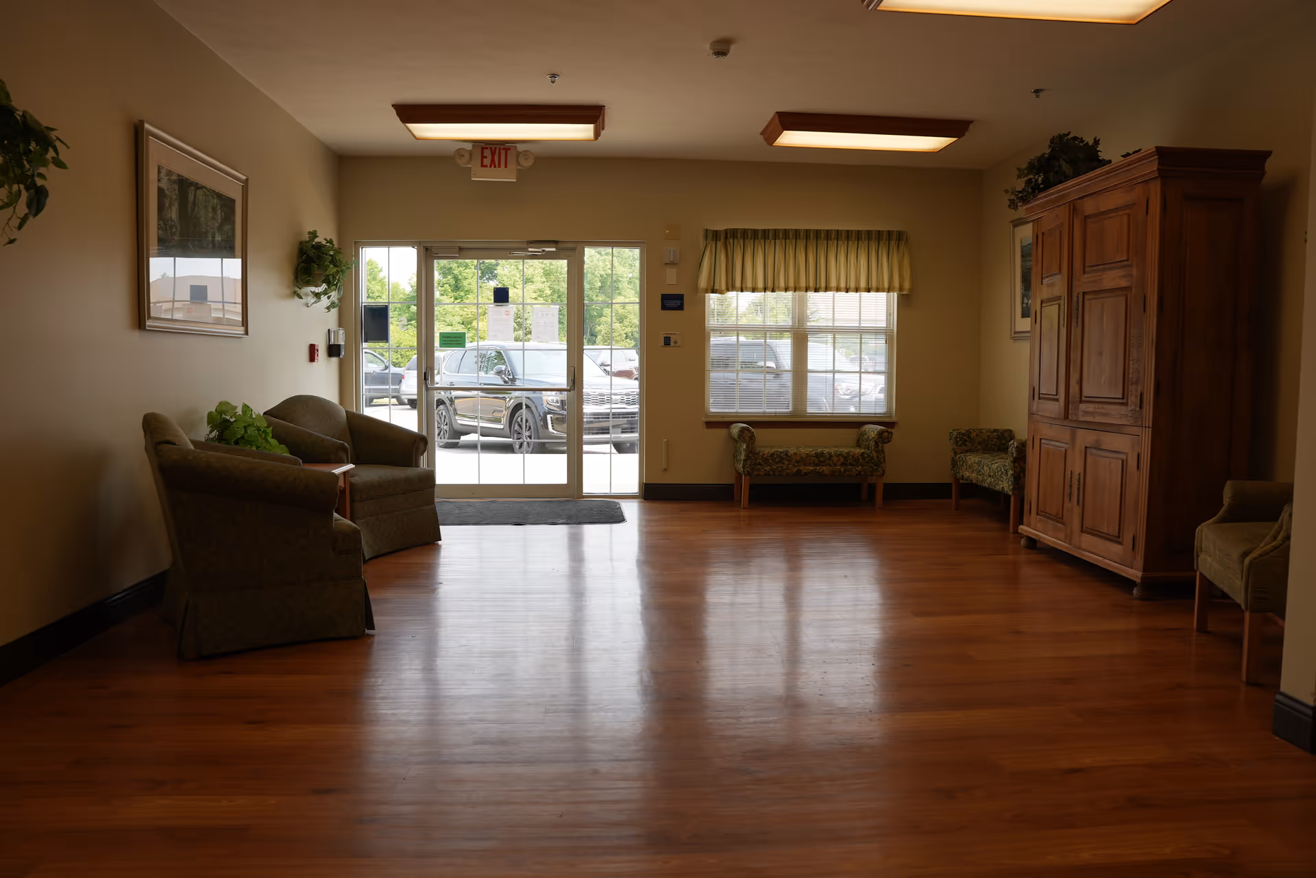 A spacious lobby with hardwood floors, upholstered chairs and benches, a large wooden cabinet, and glass exit doors showing cars outside.