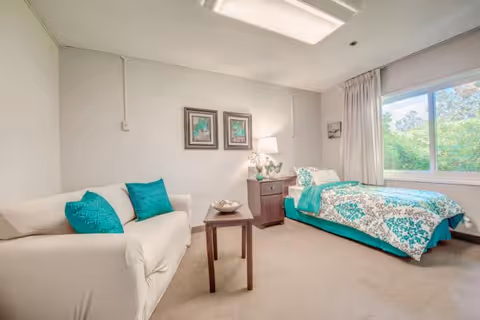 A cozy senior living bedroom featuring a single bed with teal and white patterned bedding, a white sofa with teal pillows, a wooden side table with a lamp and decorative items, two framed pictures on the wall, and a window with a view of greenery outside.
