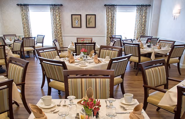 Dining room with multiple round tables set for service, striped upholstered chairs, and floral centerpieces.