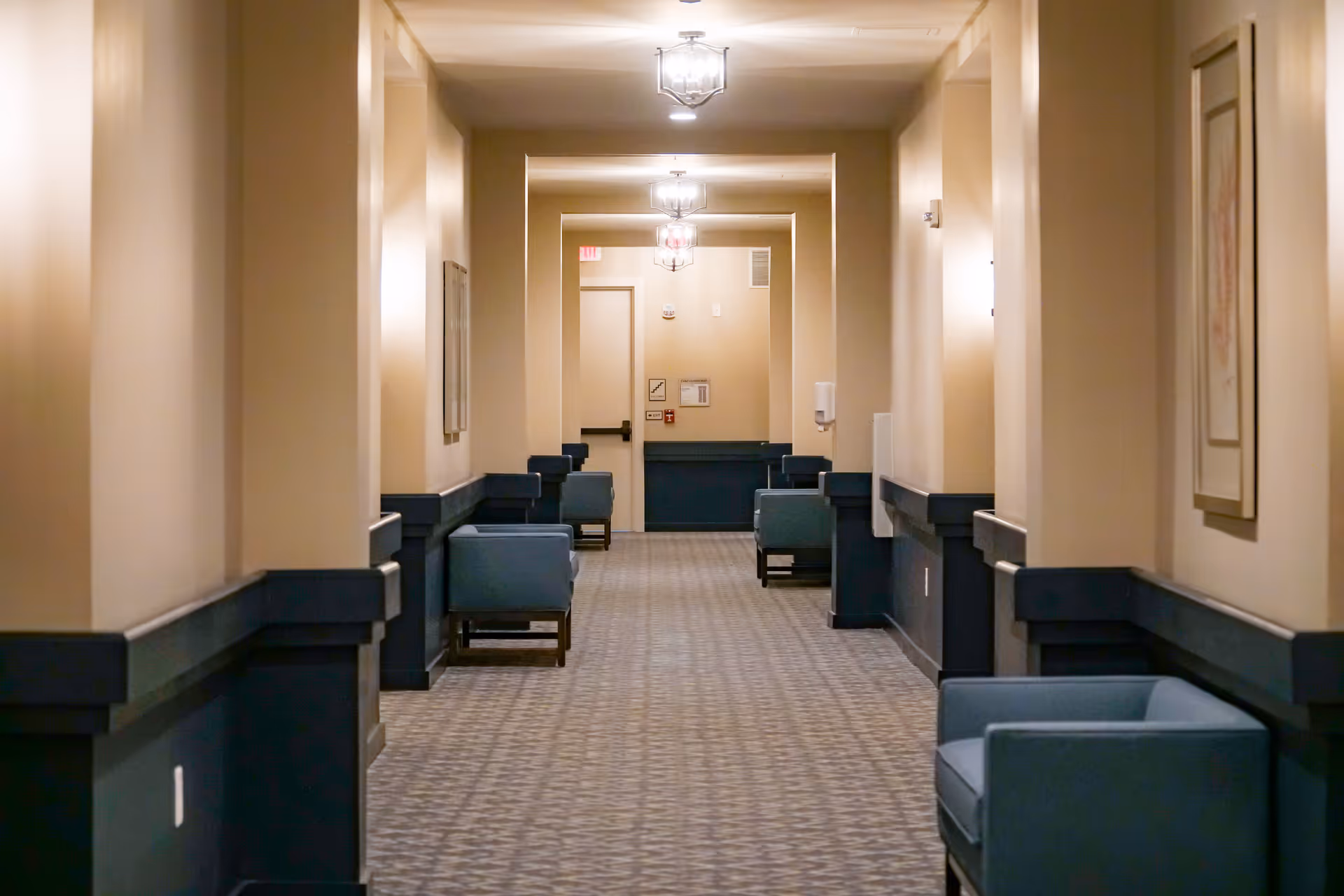 A long, well-lit hallway in a senior living facility with beige walls and dark blue wainscoting. Several blue armchairs are placed along the walls, and framed artwork hangs on the walls. Ceiling lights illuminate the corridor, and a door is visible at the end of the hallway.