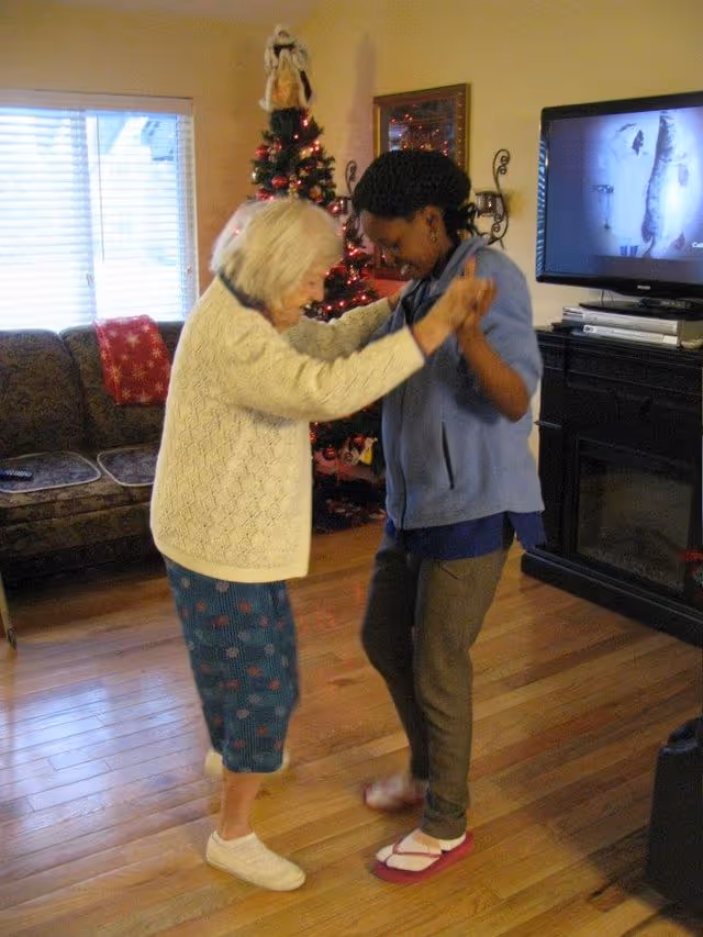 An elderly woman and a younger woman holding hands and dancing together in a living room decorated with a Christmas tree. The room has wooden floors, a couch with a red blanket, a TV on a stand, and a window with blinds.