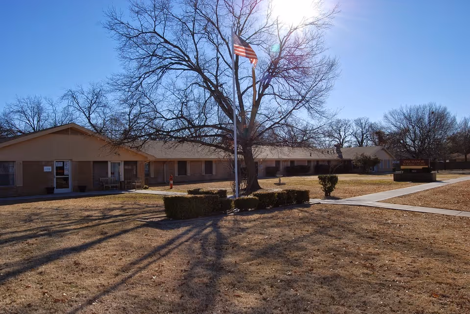 Exterior view of Oak Manor of Commerce Nursing & Rehabilitation building on a sunny day with a large leafless tree and an American flag in front. The building is single-story with a brick facade and a sign near the sidewalk. The lawn is dry with trimmed bushes and concrete walkways.