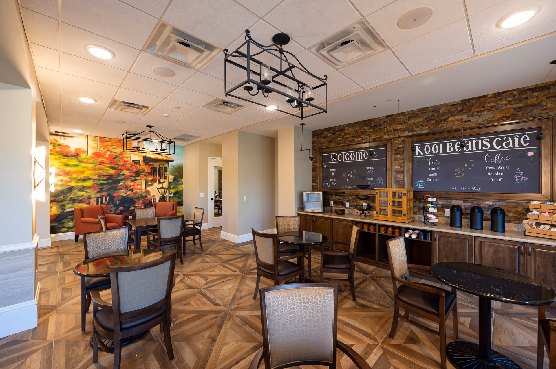 Interior view of a senior living facility café area with several tables and chairs, a mural of a colorful garden scene on one wall, and a counter with coffee and tea supplies under two chalkboard signs reading 'Welcome' and 'Kooi Beans Café'.