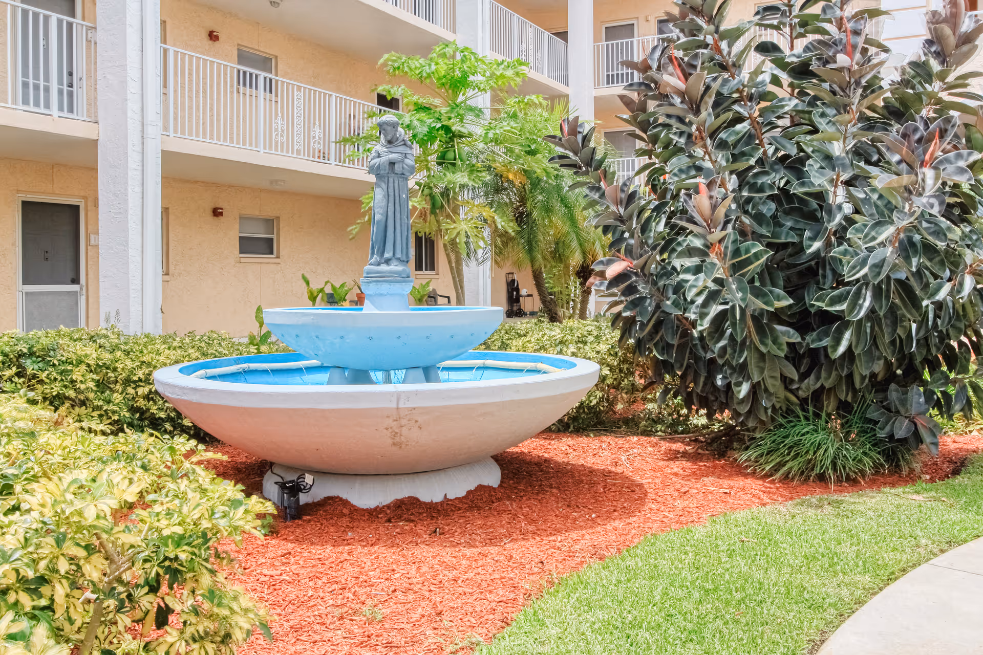 A courtyard fountain with a small statue surrounded by mulch and plants in front of a multi-story residential building.