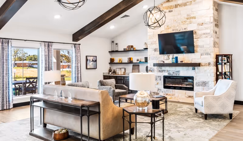 Bright spacious living room with exposed wooden beams, sofas and armchairs arranged around a stone fireplace with a mounted TV and large windows.