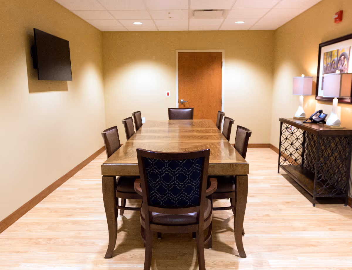 A small meeting/dining room with a long wooden table surrounded by chairs, a wall-mounted TV on the left, and a sideboard with lamps and a phone on the right.