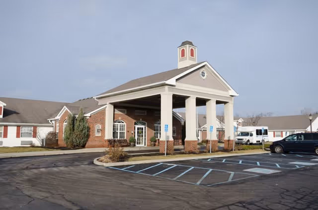 Exterior view of a senior living facility with a covered entrance supported by large white columns. The building has a combination of brick and light-colored siding with multiple windows. There are several handicap parking spaces in front, a white shuttle bus, and a dark-colored car parked nearby under a clear sky.
