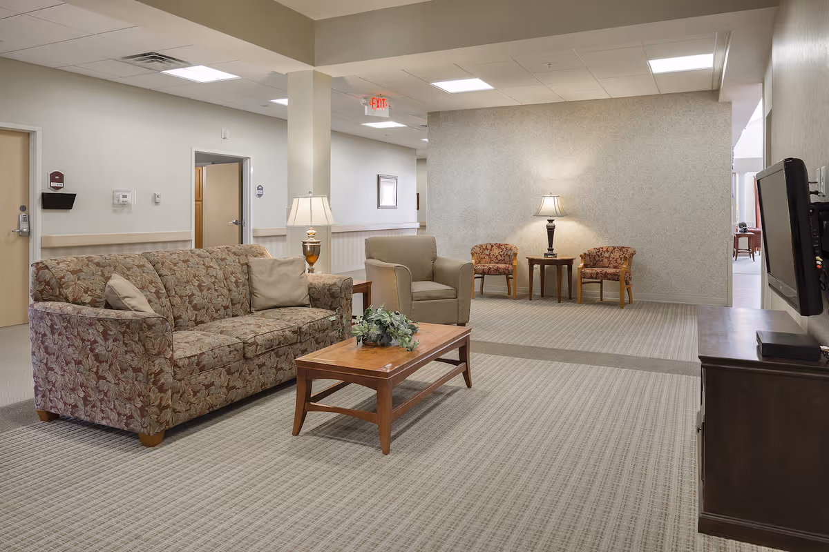 A senior living facility common area with a floral patterned sofa, a beige armchair, a wooden coffee table with a plant centerpiece, two floral armchairs with a small table and lamp between them, and a wall-mounted TV on the right side. The room has beige walls, carpeted floor, and ceiling lights.