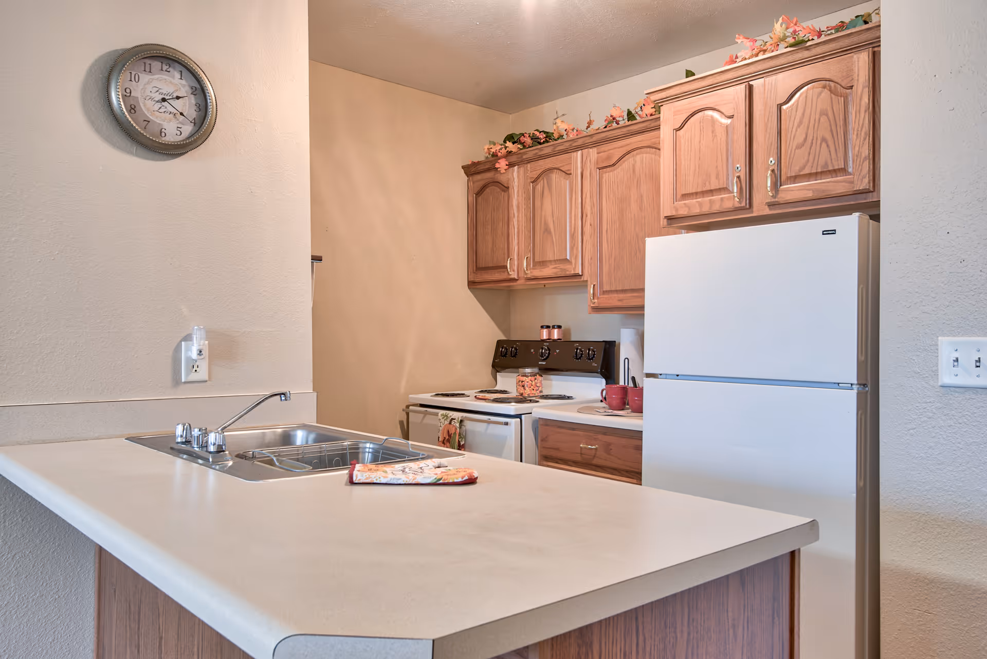 A kitchen area with wooden cabinets, a white refrigerator, a white stove with oven, and a countertop with a sink. There is a wall clock above the sink and decorative flowers placed on top of the cabinets.