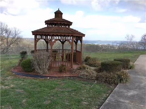 Wooden gazebo in a landscaped outdoor area with shrubs and a concrete walkway.