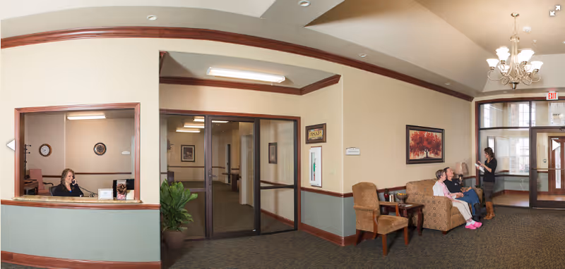 A reception area in a senior living facility with a woman sitting behind a counter on the left, a hallway with glass doors in the center, and a seating area on the right where two elderly people are sitting on a couch and a woman is standing and talking to them. The room has beige walls with wood trim, a chandelier, and framed artwork on the walls.