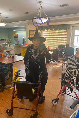 An elderly woman dressed in a witch costume with a black hat and tinsel garland stands with a walker in a common room. Other elderly individuals are seated nearby, some in wheelchairs or using walkers. The room has wooden floors, tables, chairs, and large windows with blinds.