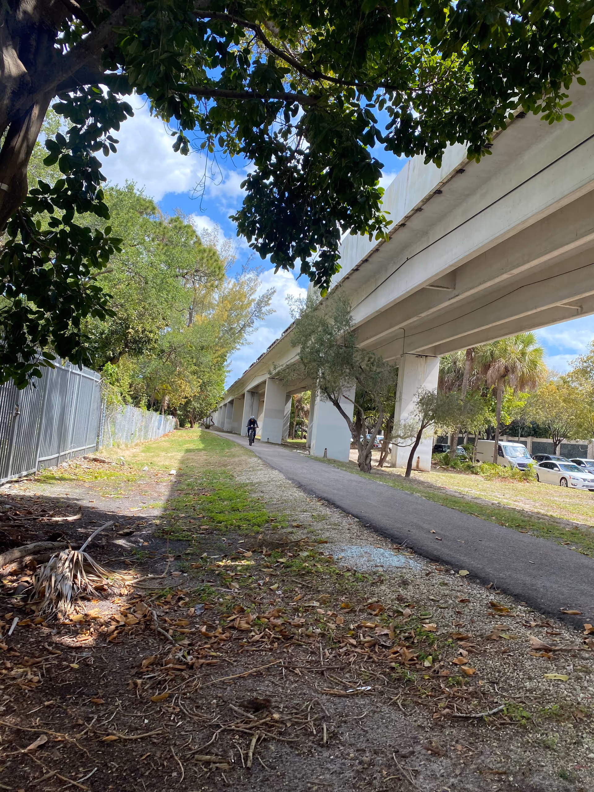 A paved pathway running alongside a large elevated concrete structure, possibly a bridge or train track, with trees and greenery on both sides. A person is seen walking or biking on the path. There are parked cars visible in the background under the structure. The sky is partly cloudy with patches of blue.