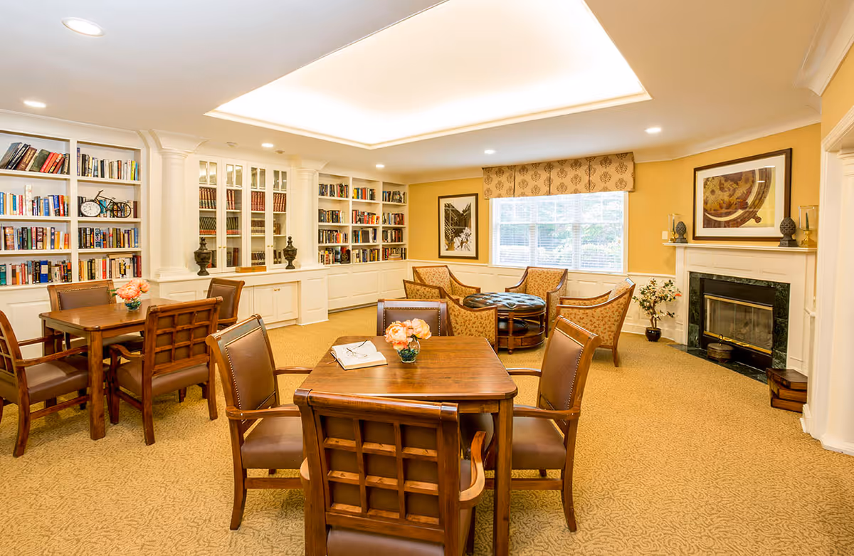 A cozy senior living common area with wooden tables and chairs, a built-in bookshelf filled with books, a fireplace with decorative items on the mantel, and a seating area with upholstered armchairs and a round ottoman near a window with patterned valance.