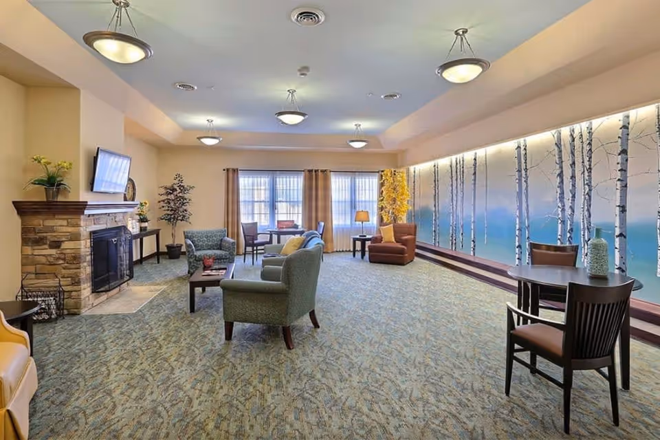 Bright communal lounge with multiple seating areas, a stone fireplace, tables, and a birch-tree mural along one wall.